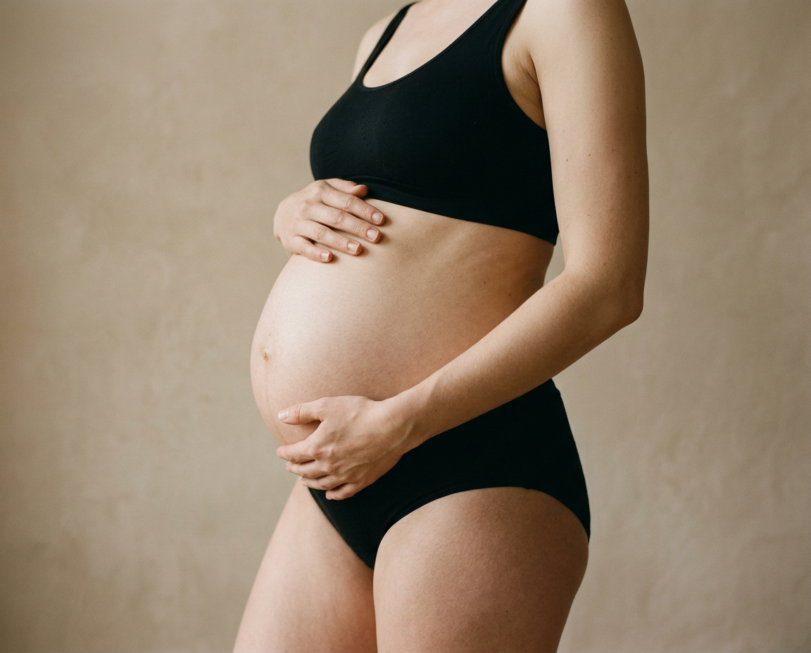 Photographie éditoriale sobre d'une femme enceinte de huit mois en sous-vêtements noirs tenant son ventre, cadrée sans le visage